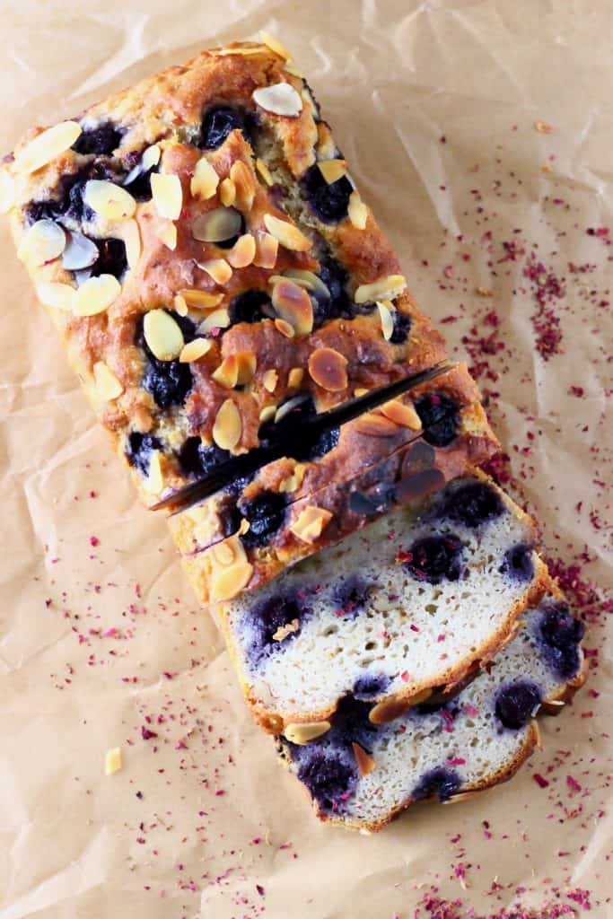 A loaf of blueberry banana bread with three slices next to it on a sheet of brown baking paper