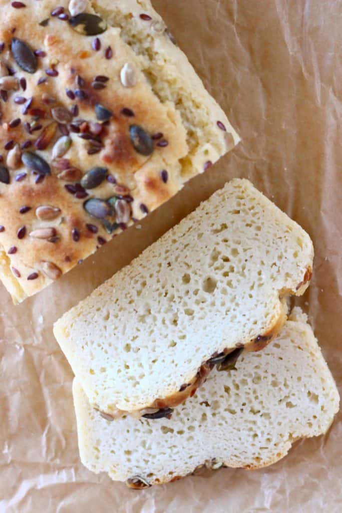 A loaf of brown bread sprinkled with mixed seeds with two slices of white bread next to it against a sheet of brown baking paper