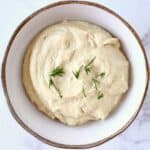 Vegan cream cheese topped with sprigs of rosemary in a white bowl with a brown rim against a marble background