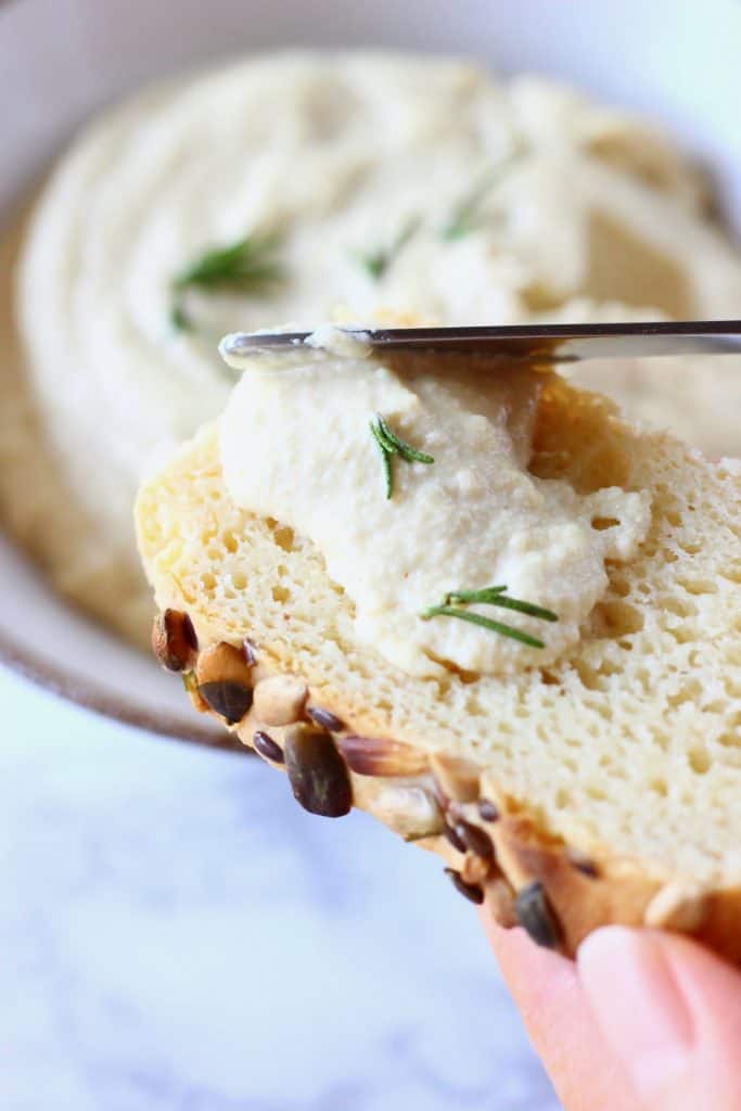 Vegan cream cheese in a bowl topped with sprigs of rosemary with a silver knife spreading a bit of it on a slice of white bread