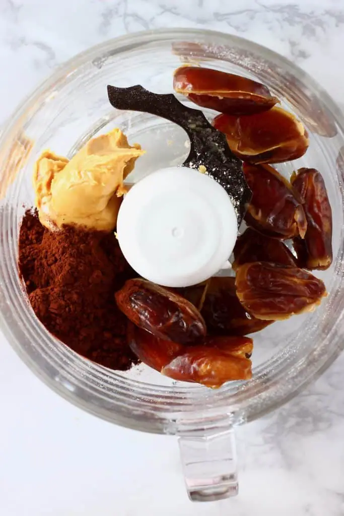 Dates, peanut butter and cocoa powder in a food processor against a marble background