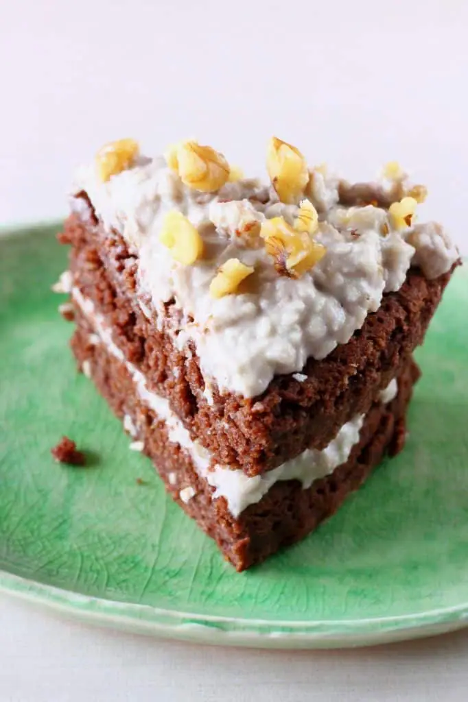 A slice of chocolate cake topped with white frosting and walnuts on a green plate against a white background
