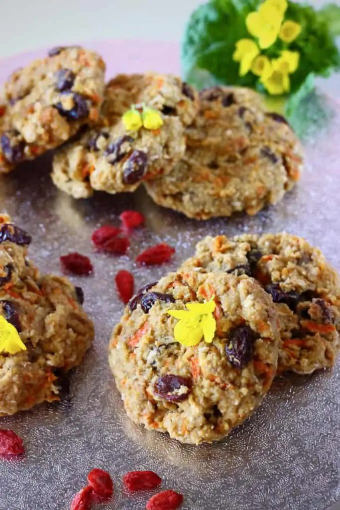 Several carrot cake cookies with raisins on a silver surface sprinkled with goji berries and yellow flowers