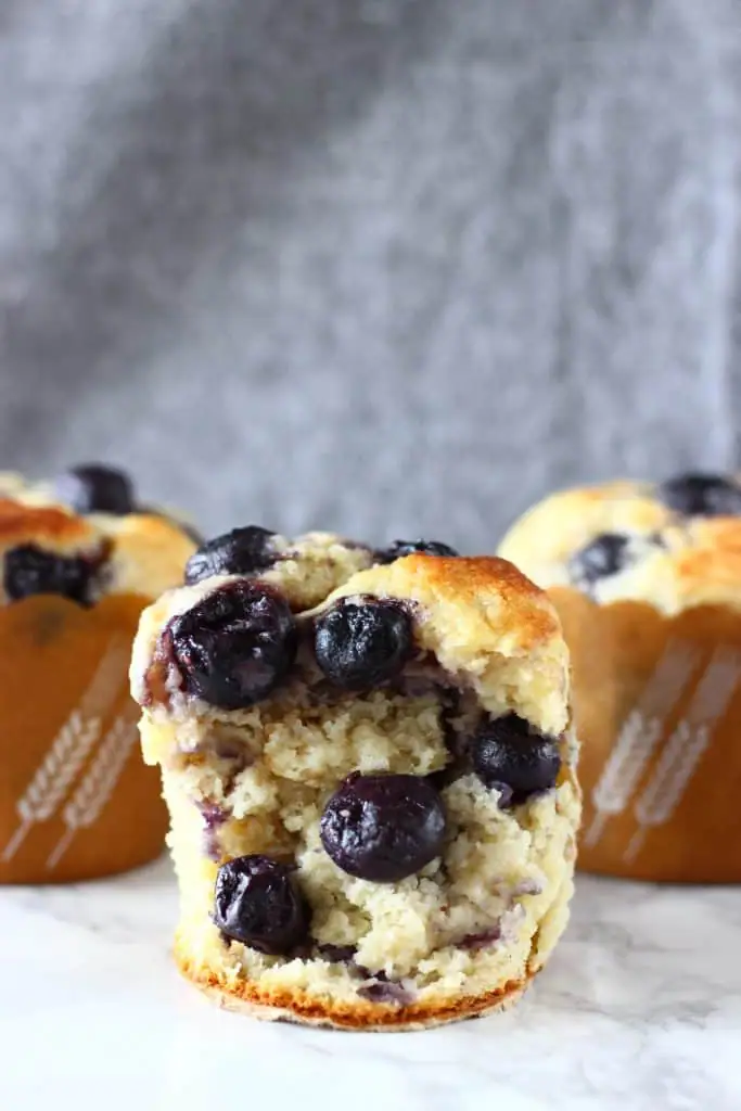 A blueberry muffin with two more muffins in the background on a marble surface against a grey background