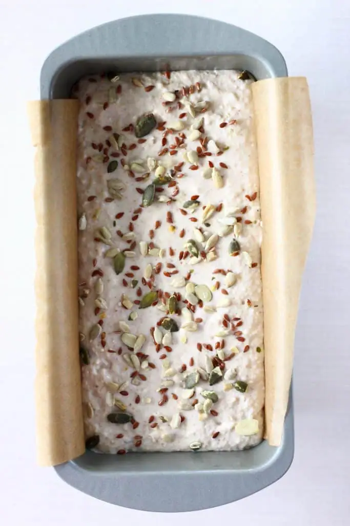 White bread batter scattered with mixed seeds in a silver loaf tin lined with brown baking paper against a white background