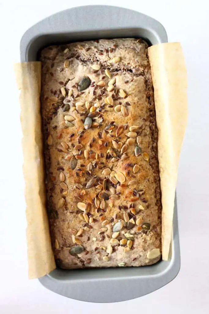 A loaf of golden brown bread topped with mixed seeds in a silver loaf tin lined with brown baking paper against a white background