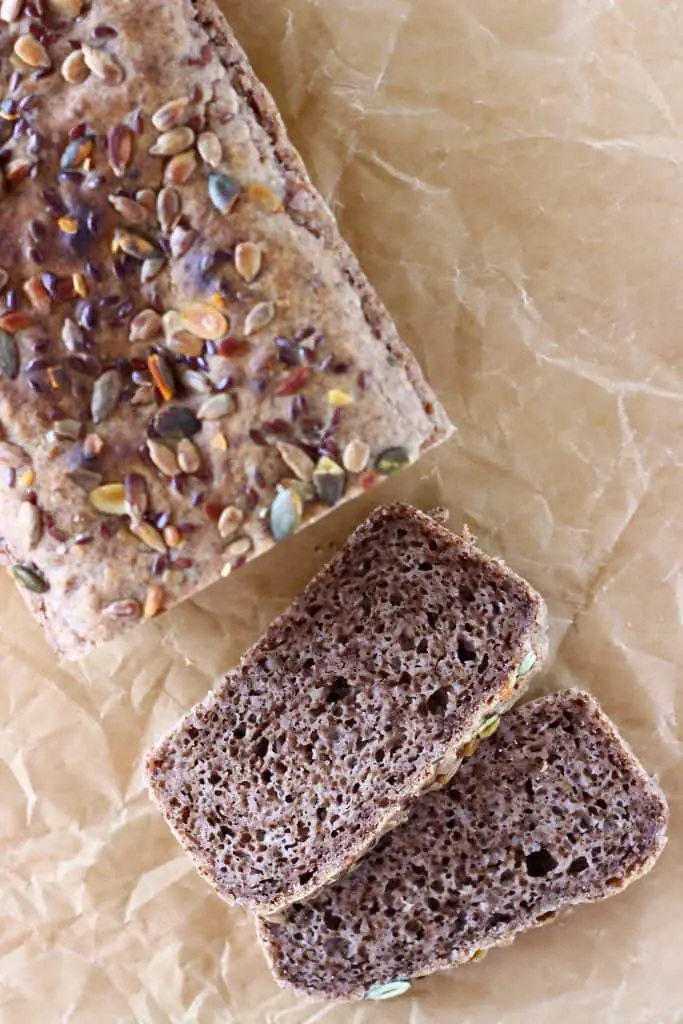 A loaf of brown bread topped with mixed seeds with two slices next to it against a sheet of brown baking paper