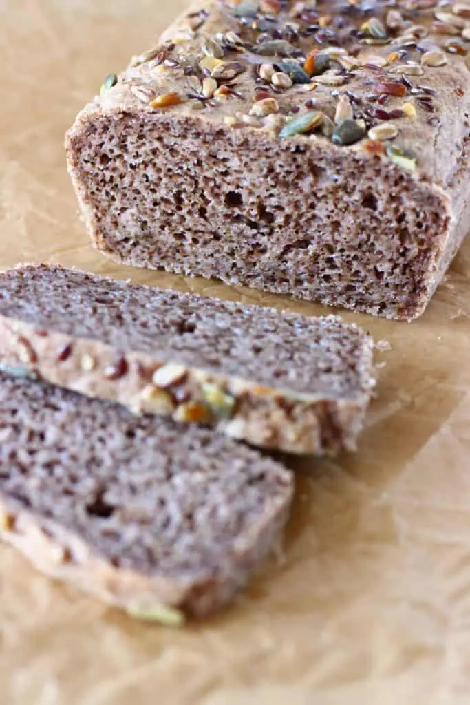 A loaf of brown bread topped with mixed seeds with two slices next to it against a sheet of brown baking paper