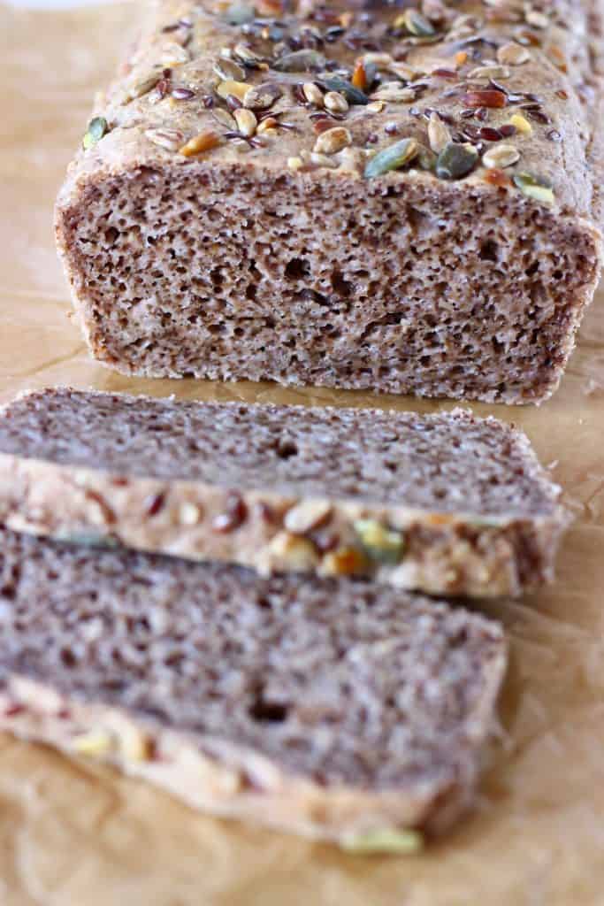 A loaf of brown bread topped with mixed seeds with two slices next to it against a sheet of brown baking paper