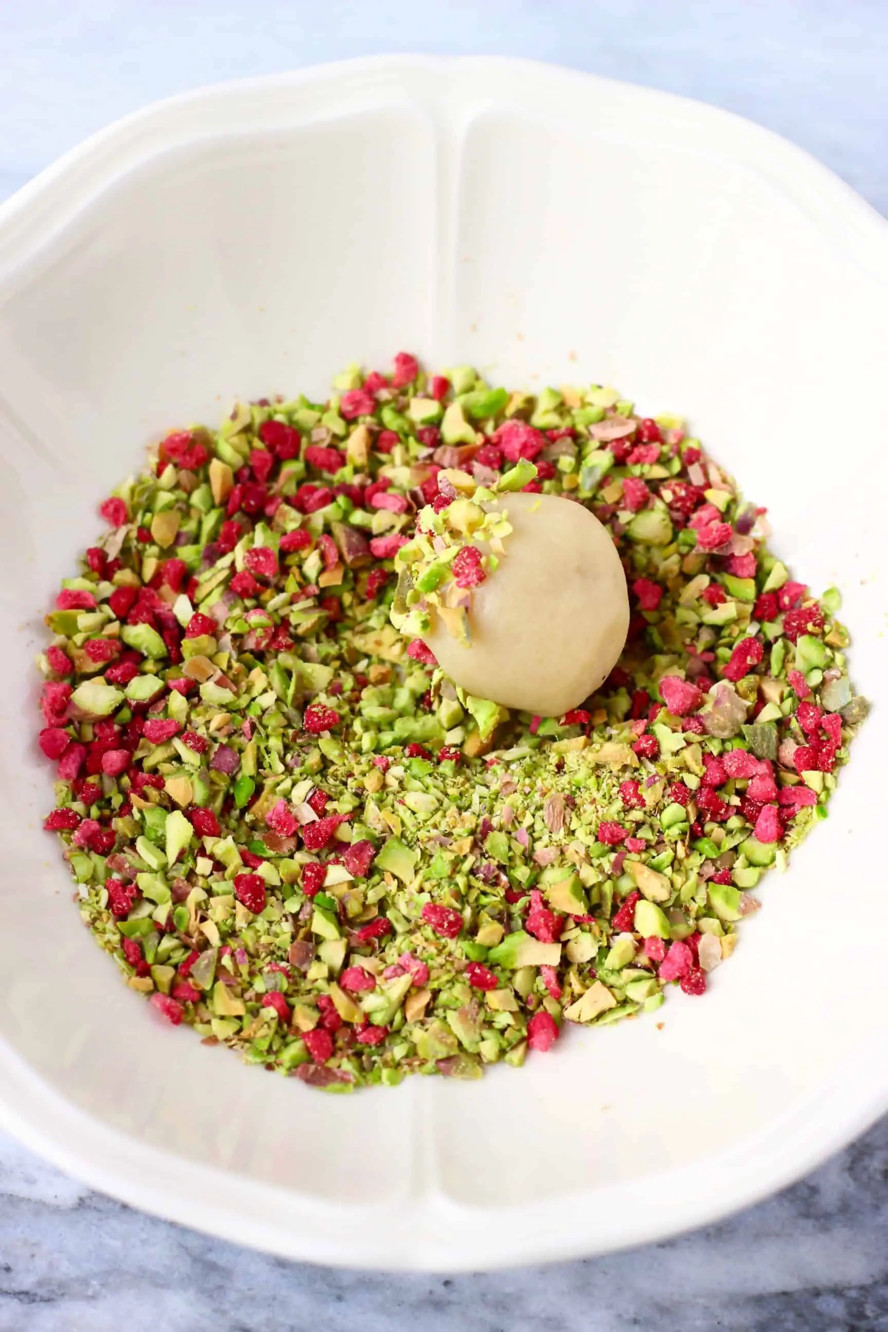A vegan white chocolate truffle being rolled around in a bowl of chopped pistachios and freeze-dried raspberries 