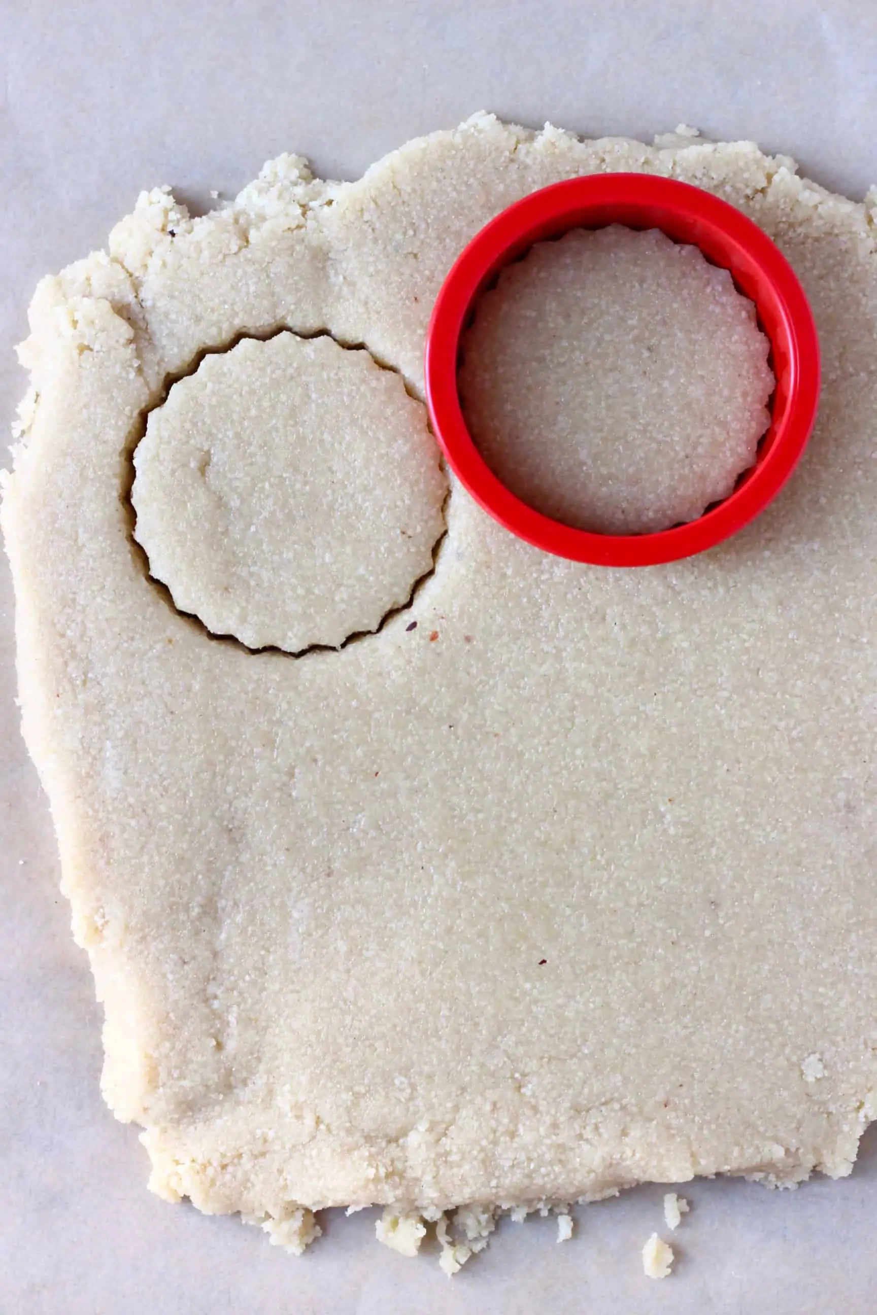 A square of raw vegan shortbread cookie dough rolled out on baking paper with a circular cookie cutter cutting out a circle of dough