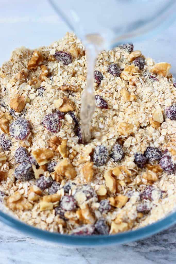Oats, dried cranberries and chopped walnuts in a glass mixing bowl with a jug of water being poured into it