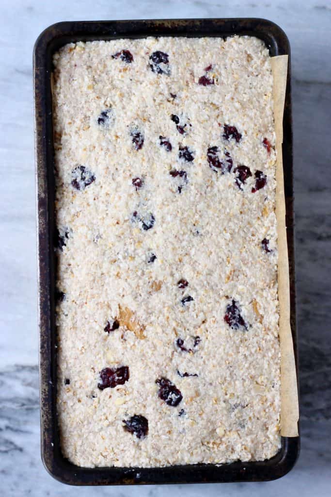 Raw oatmeal bread batter with cranberries and chopped walnuts in a black loaf tin against a marble background
