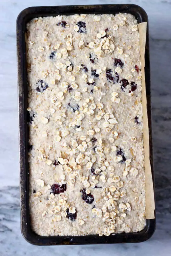 Raw oatmeal bread batter with cranberries and chopped walnuts sprinkled with oats in a black loaf tin against a marble background