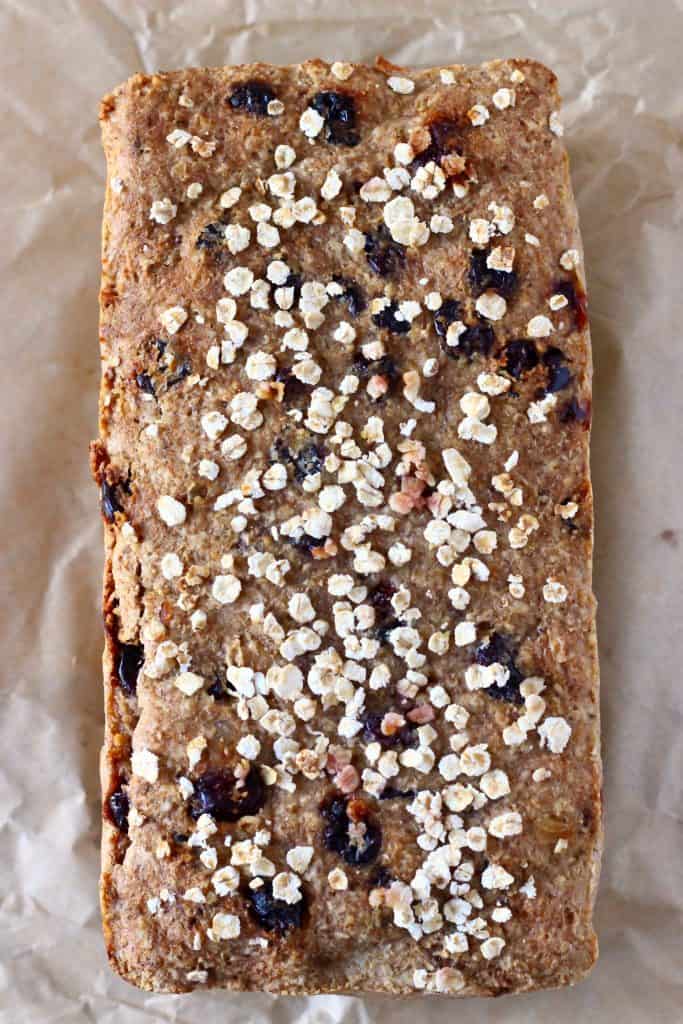 A loaf of brown bread with cranberries sprinkled with oats on a sheet of brown baking paper