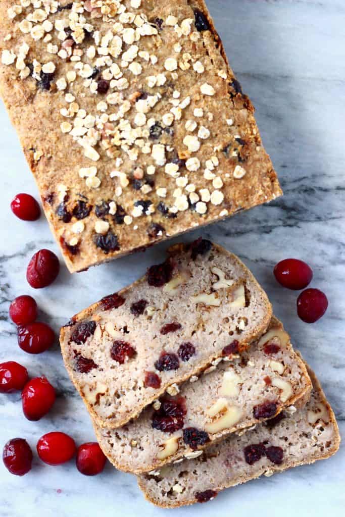 A loaf of brown bread with cranberries sprinkled with oats with three slices cut from it on a marble background scattered with fresh cranberries