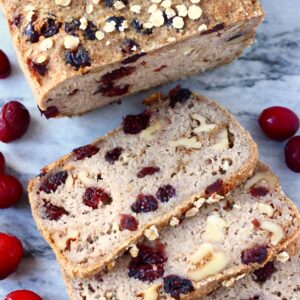 A loaf of brown bread with cranberries sprinkled with oats with three slices cut from it on a marble background scattered with fresh cranberries