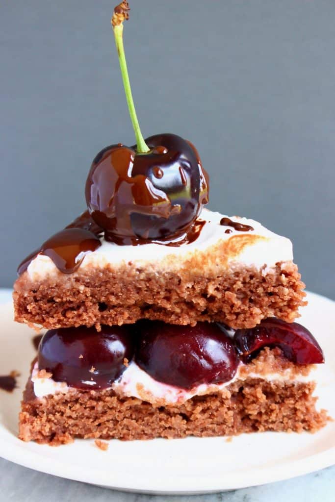 Photo of chocolate sponge cake sandwiched with white creamy frosting and red cherries with a whole cherry on top against a grey background