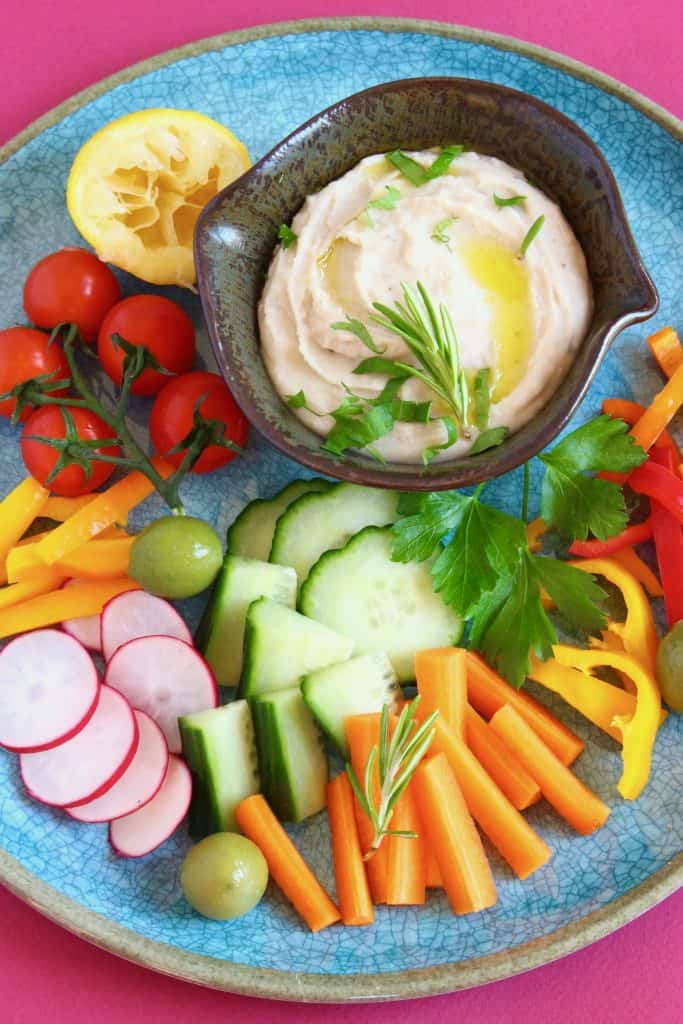 Photo of a black bowl filled with white hummus decorated with green herbs on a blue plate with chopped vegetables