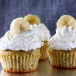 Three banana cupcakes topped with cream and a banana slice each on a silver board against a grey background