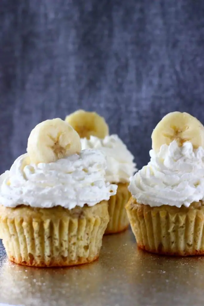 Three banana cupcakes topped with cream and a banana slice each on a silver board against a grey background