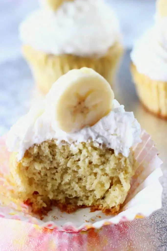 Half a cupcake topped with cream and a slice of banana in a cupcake wrapper against a silver board with two more cupcakes in the background 