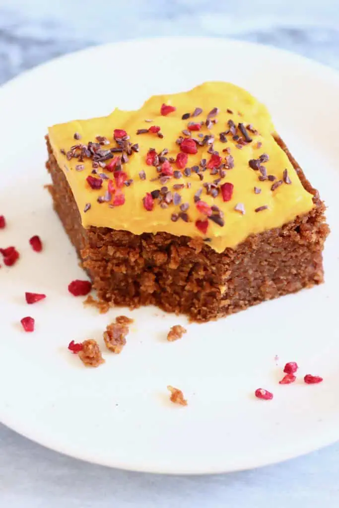 A square of chocolate sponge cake topped with yellow frosting sprinkled with cacao nibs and freeze-dried raspberries with a mouthful taken out of it on a white plate against a marble background