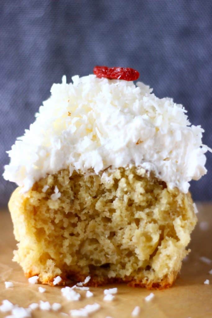 A yellow cupcake topped with white creamy frosting, desiccated coconut and two goji berries against a grey background