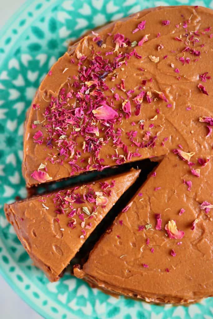 A chocolate sponge cake sprinkled with rose petals with a slice taken out of it on a green cake stand