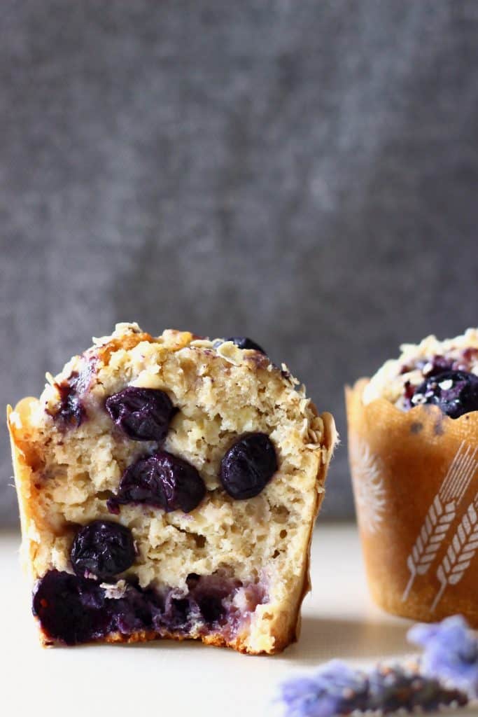 A halved blueberry muffin with a whole blueberry muffin in the background on a white surface against a grey background