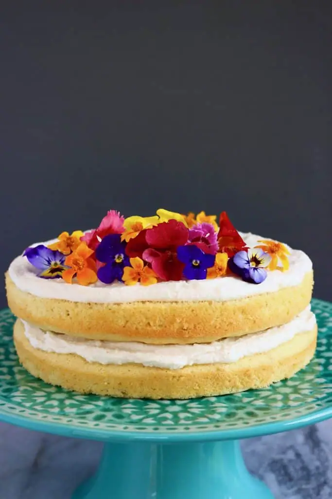A sponge cake layered with white creamy frosting and topped with colourful edible flowers on a green cake stand against a grey background