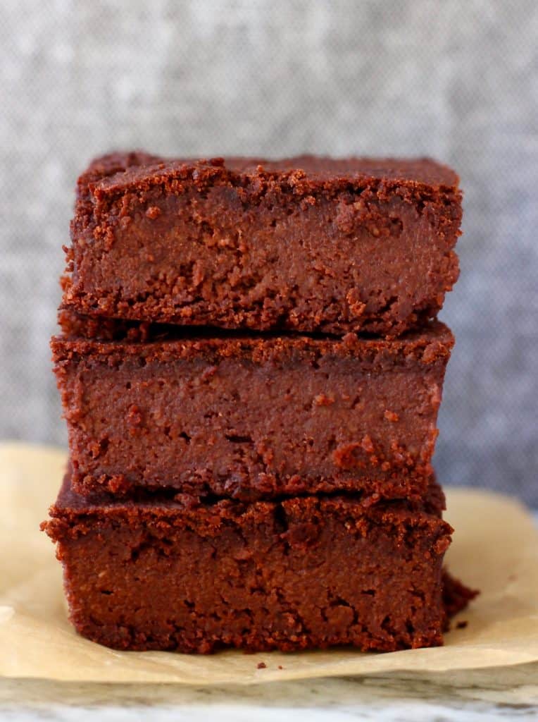 Photo of three chocolate brownies stacked on top of each other on a piece of brown baking paper against a grey background