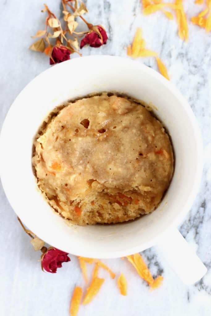 An orange mug cake in a white mug against a marble background with dried roses