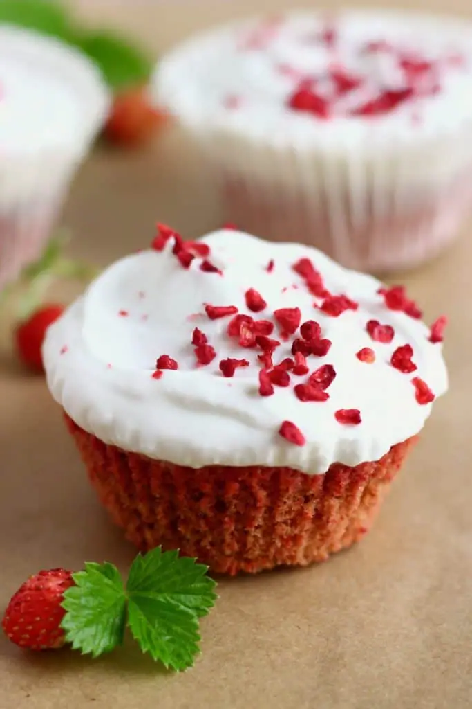 A red velvet cupcake topped with white creamy frosting and freeze dried raspberries with two more cupcakes in the background