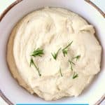Vegan cream cheese topped with sprigs of rosemary in a white bowl with a brown rim against a marble background