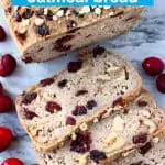 A loaf of brown bread with cranberries sprinkled with oats with three slices cut from it on a marble background scattered with fresh cranberries