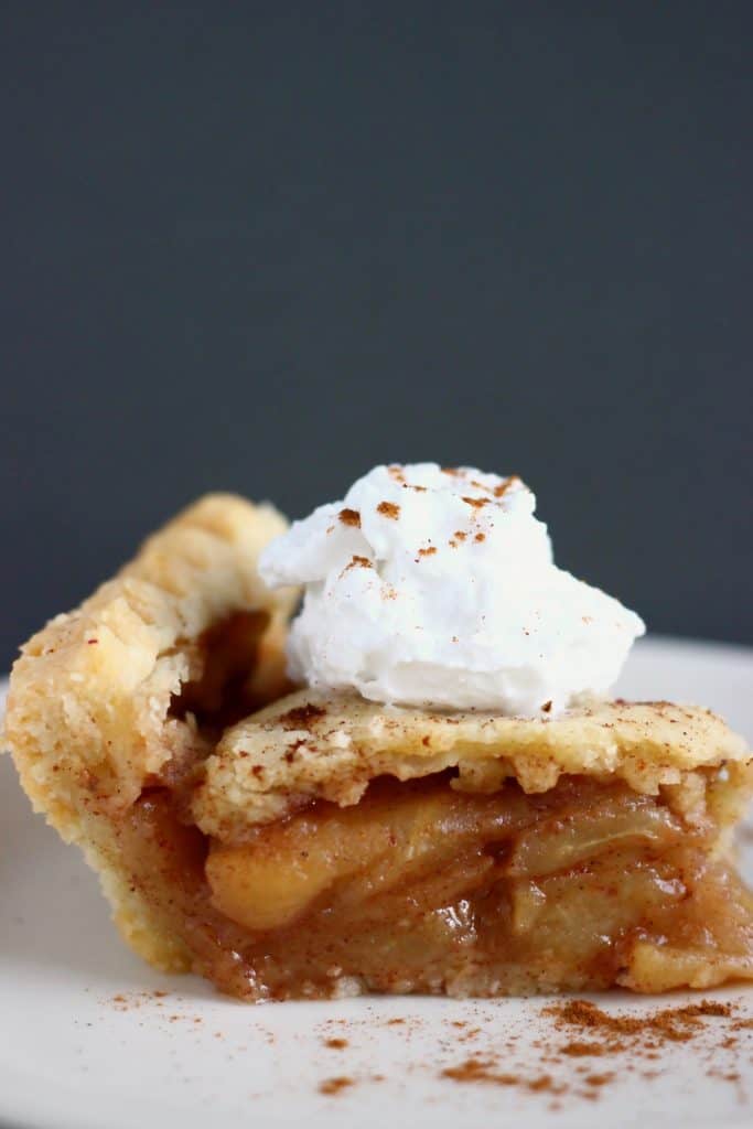 A slice of apple pie topped with cream on a white plate sprinkled with cinnamon against a grey background