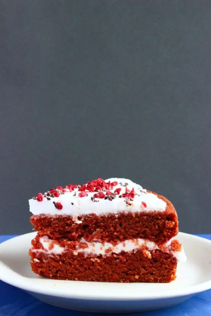 A slice of red velvet cake with white creamy frosting sprinkled with freeze-dried raspberries on a white plate against a grey background