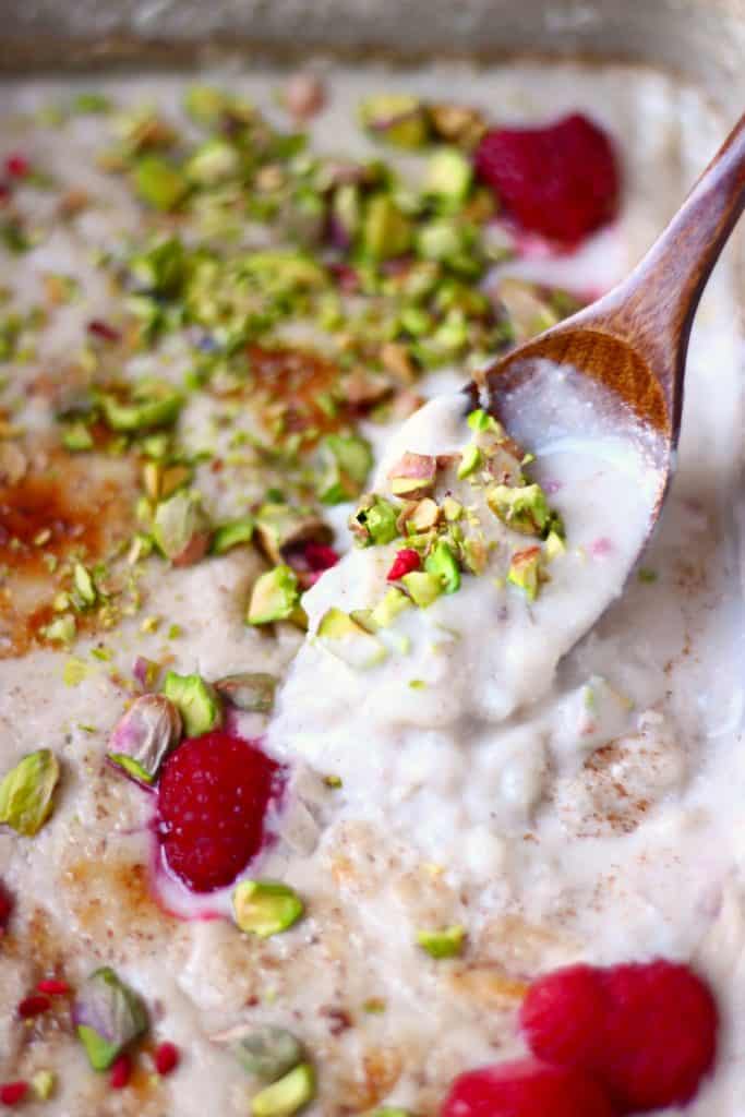 Rice pudding in a grey rectangular baking dish topped with chopped pistachios and fresh raspberries with a wooden spoon lifting up a mouthful of it