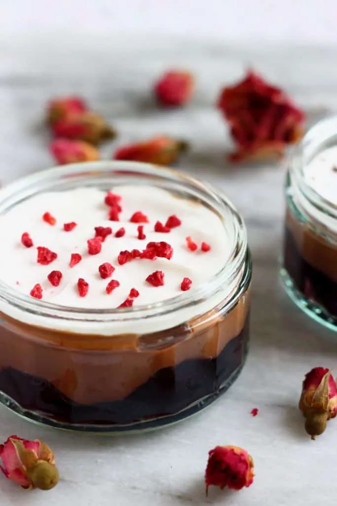 Photo of a trifle in a glass ramekin made with cherries, chocolate custard and topped with white cream and freeze-dried raspberries against a marble background scattered with dried roses