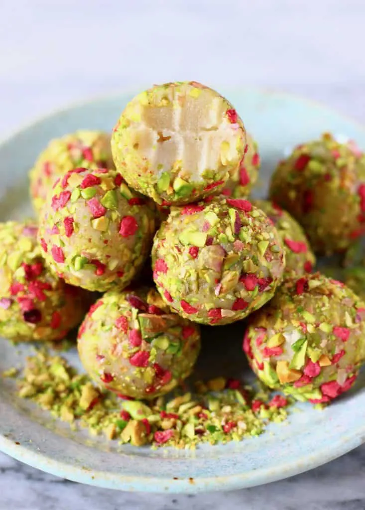 A pile of white chocolate truffles covered in chopped pistachios and freeze-dried raspberries with a bitten one on top on a light blue plate against a marble background