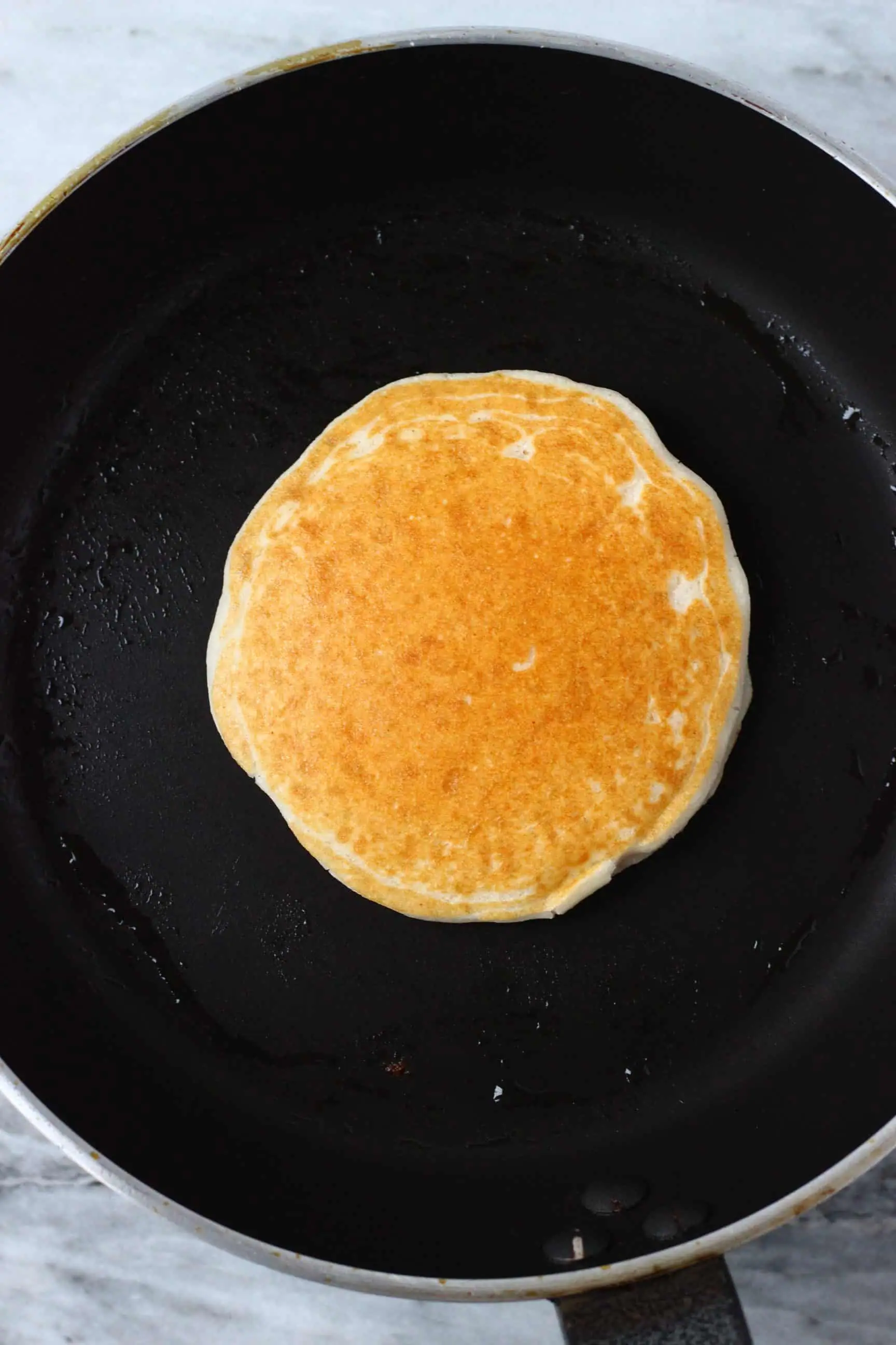 A golden brown vegan coconut flour pancake being cooked in a black frying pan 