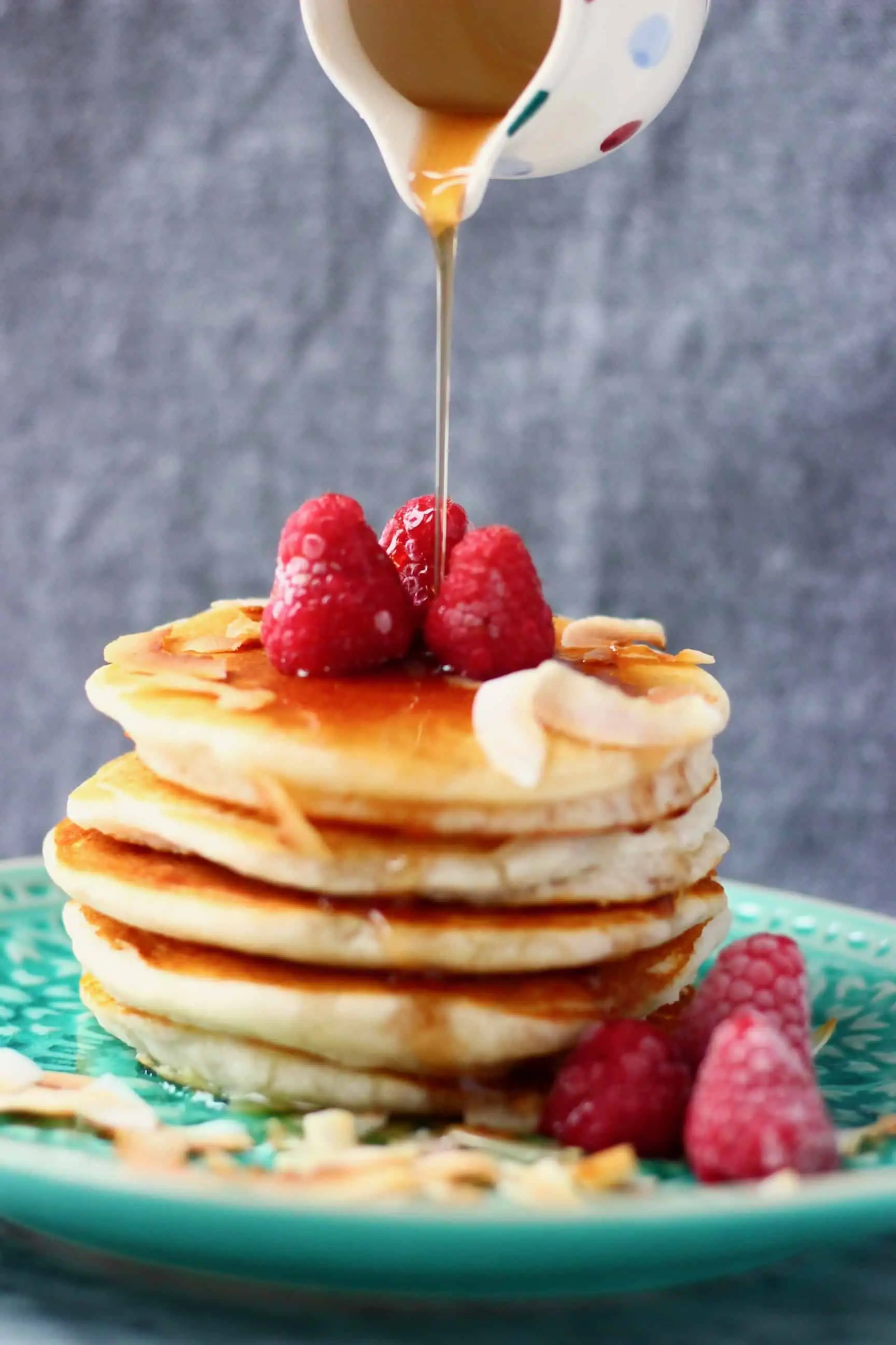 Five vegan coconut flour pancakes stacked in a pile decorated with fresh raspberries with a small jug pouring syrup over the top 