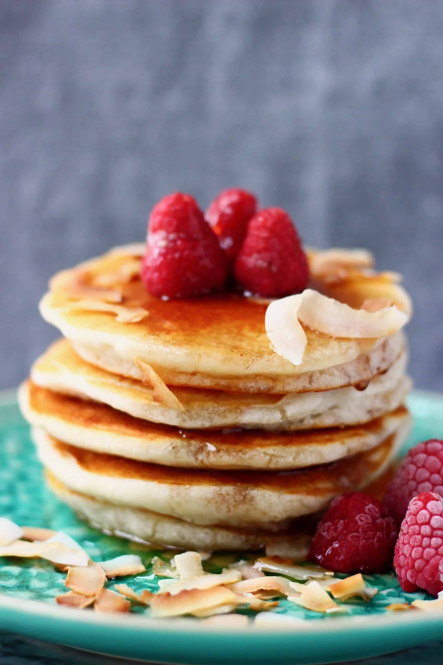 Five vegan coconut flour pancakes stacked in a pile decorated with fresh raspberries on a green plate against a grey background