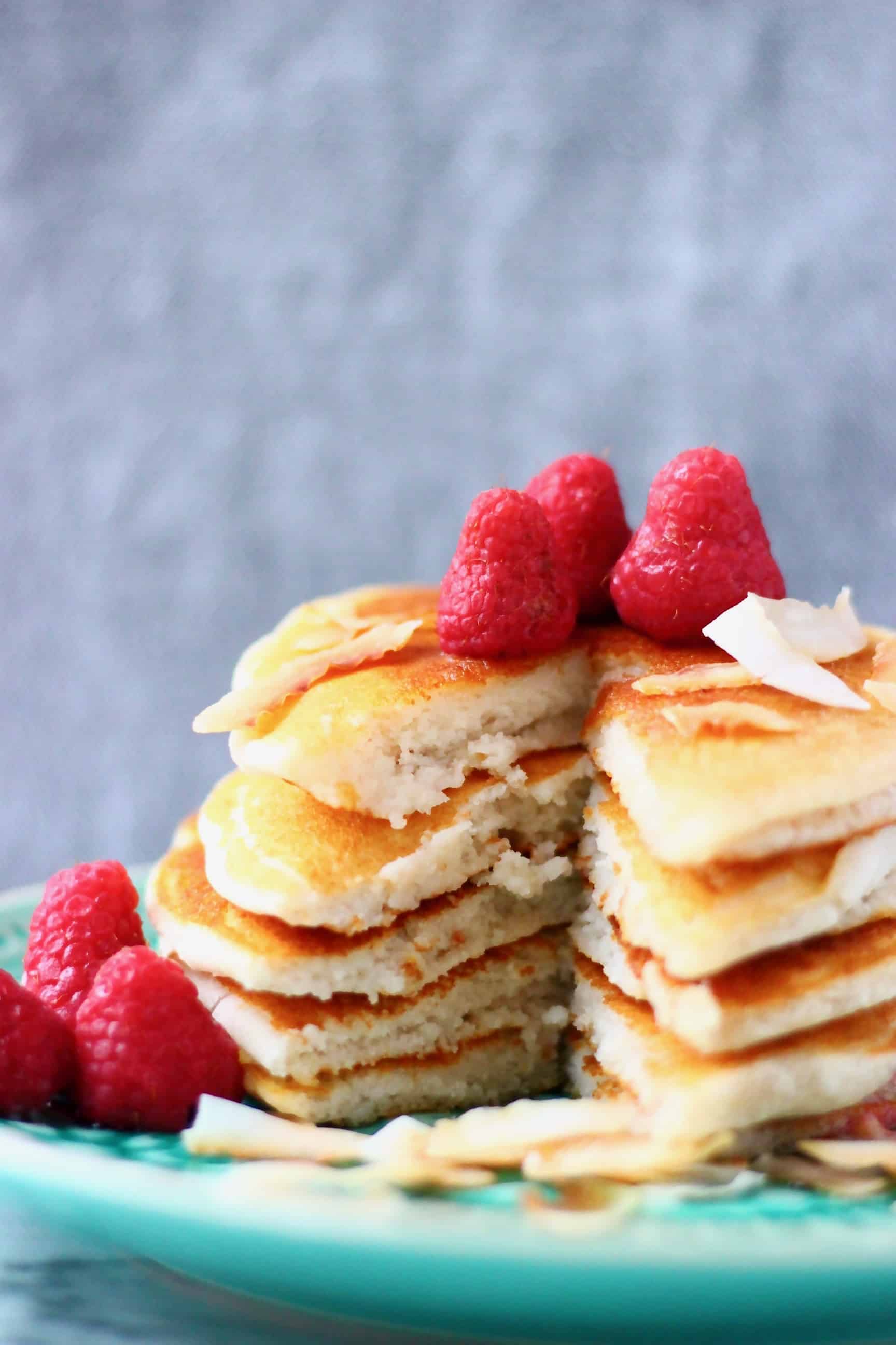 Five coconut flour pancakes stacked in a pile decorated with fresh raspberries and coconut flakes on a green plate against a grey background