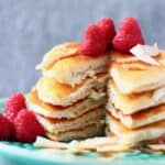 Five vegan coconut flour pancakes stacked in a pile decorated with fresh raspberries and coconut flakes on a green plate against a grey background