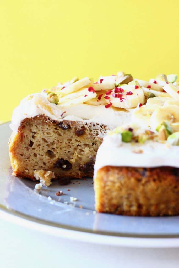 A sliced banana cake on a green plate topped with white frosting, banana slices and chopped pistachios against a yellow background