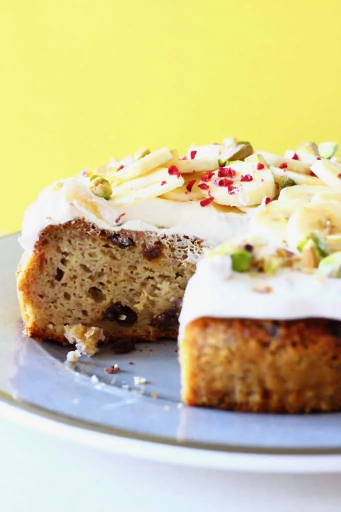 A sliced banana cake on a green plate topped with white frosting, banana slices and chopped pistachios against a yellow background