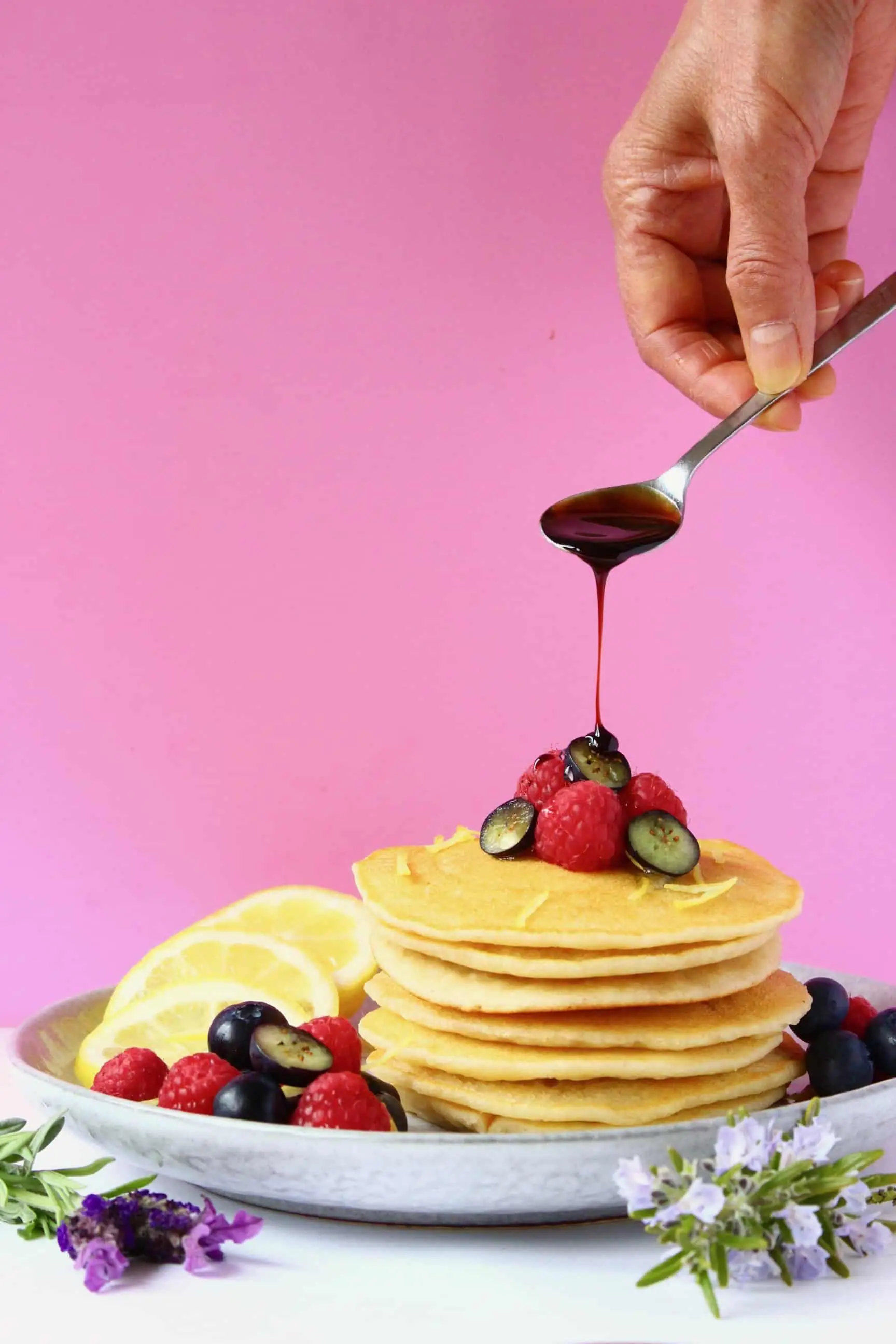 A stack of gluten-free vegan pancakes topped with fresh berries with a spoon pouring syrup over them