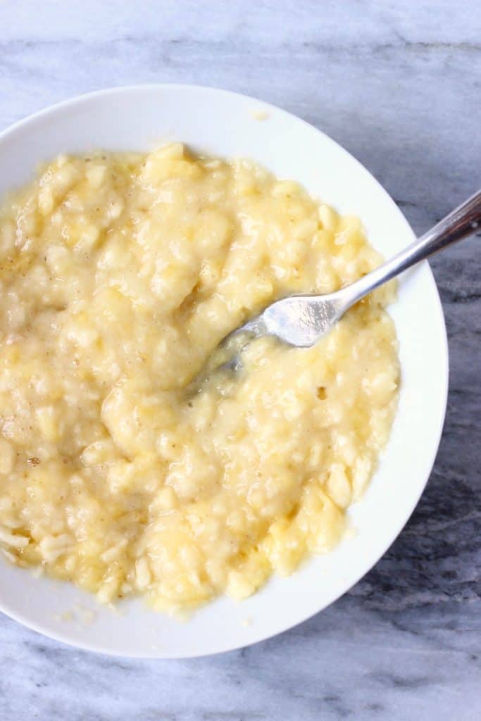 Mashed banana in a white bowl with a silver fork against a marble background